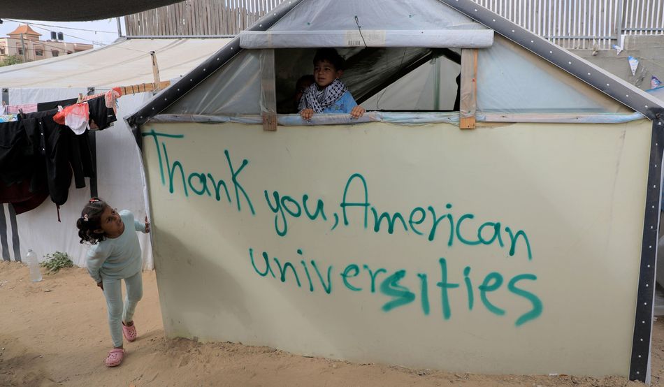 The words "Thank you, American universities" is scrawled in green on a temporary shelter in Gaza. Two children peer at it.