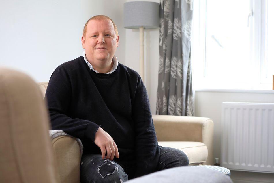 Leigh Midwinter, a white man wearing a black jumper, looks into the camera from his place on a beige sofa in a white room
