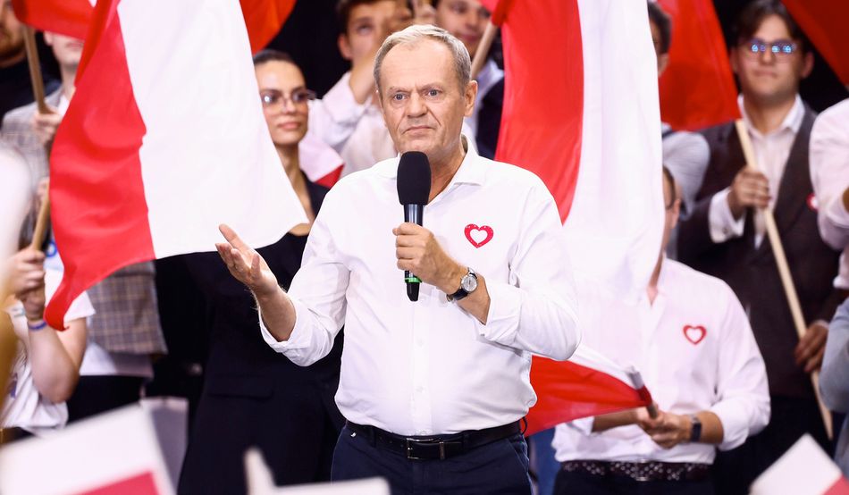 Donald Tusk, a man with blonde hair and wearing an open-necked white shirt, speaks into a handheld microphone in front of a crowd wearing black and white and waving red and white flags©Alamy