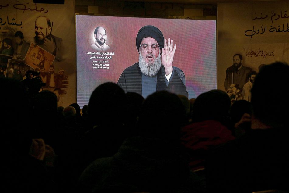 Secretary general of Hezbollah Hassan Nasrallah, a man wearing black with a grey beard, is shown raising his left hand, palm shown, on a big video screen. He is watched by a crowd of people, shown as silhouettes
