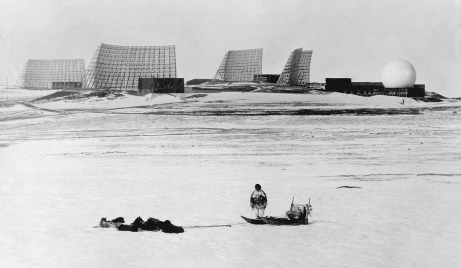 A black and white photo of a person standing next to a dogsled, their back to the camera, looking at the air base. The airbase is formed of radio towers and a white, spherical structure. Image: NF/SCANPIX/AFP via Getty Images
