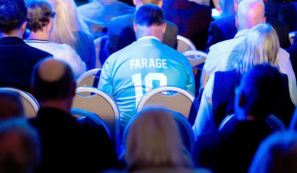 People are seated in rows, shown from behind. A man is wearing a pale blue football-style shirt with the name Farage and the number 10 on the back. Image: Alamy
