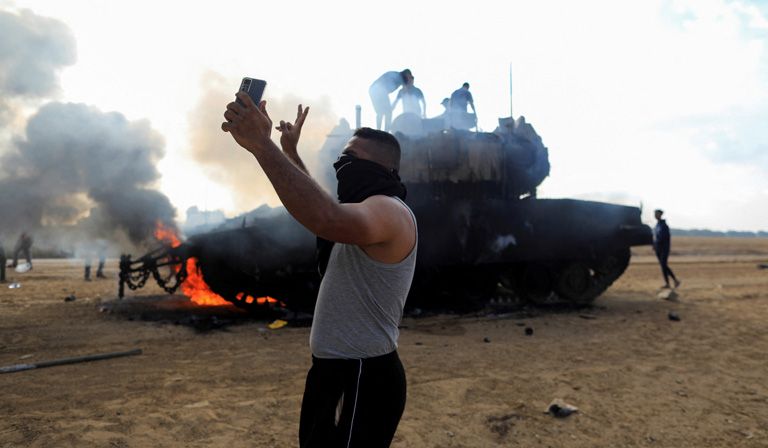 A man takes a selfie in front of a burning Israeli tank. The tank is emitting smoke and several people stand on top of it© REUTERS/Yasser Qudih