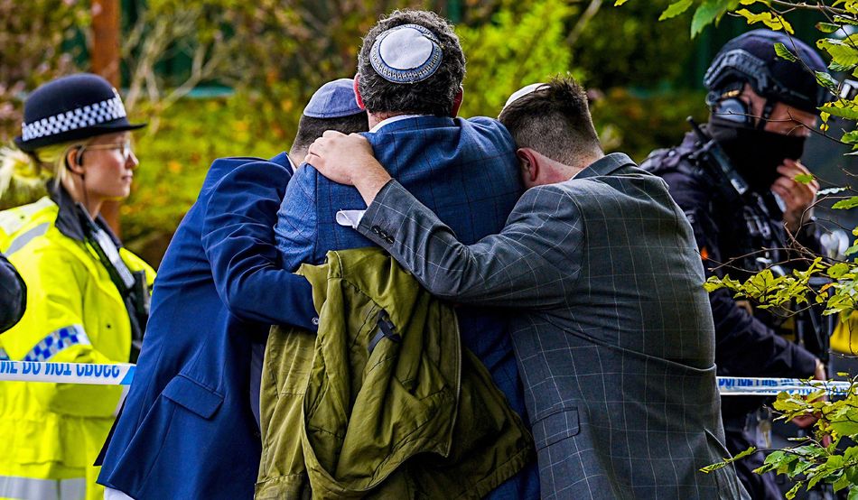 Three Jewish men, each wearing a suit and a kippah, hug one another. Nearby is a police officer in high-visibility clothing, and another armed officer dressed in all black. Image: Alamy