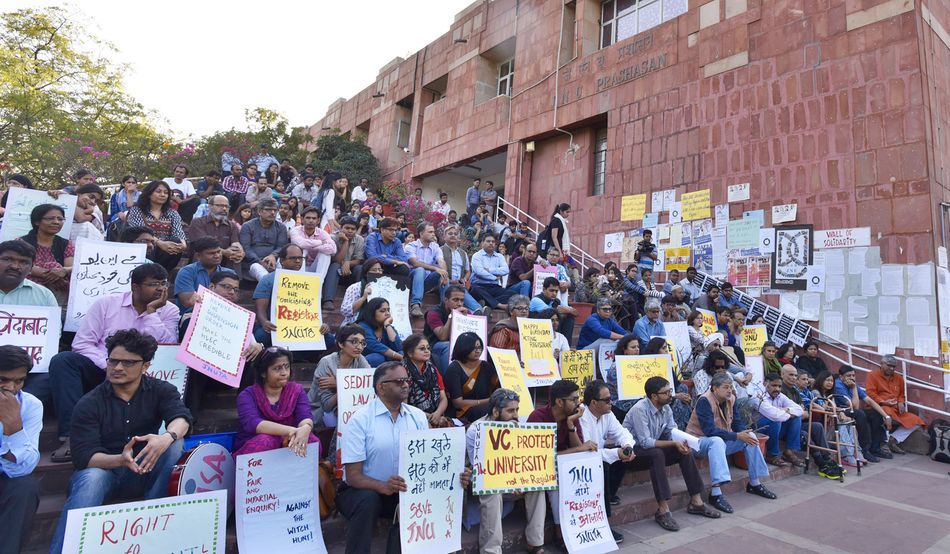 Protesters sit on steps at JNU, holding placards. The building behind them is a pinkish red. Image: Sanjeev Verma-Hindustan Times via Getty Images