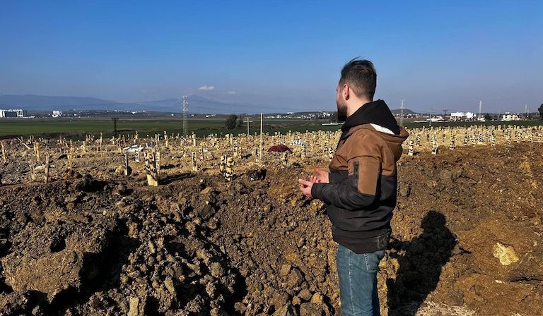 Fadi, a man with dark hair and wearing jeans and a hoodie, looks at the rough soil of a graveyard in Antakya
