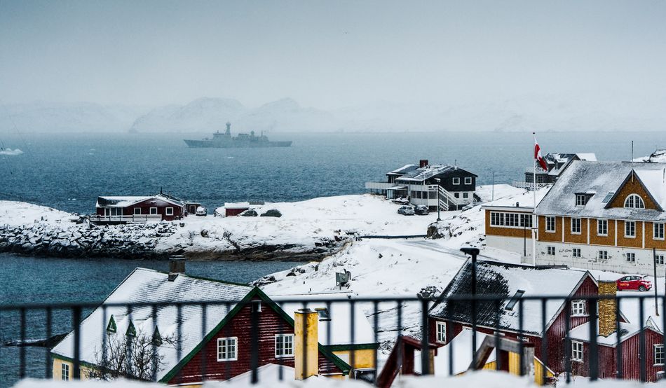 In the foreground are the snow covered red and yellow buildings of Nuuk. Behind, a Danish navy patrol ship sails through the ocean. Image: AFP via Getty Images
