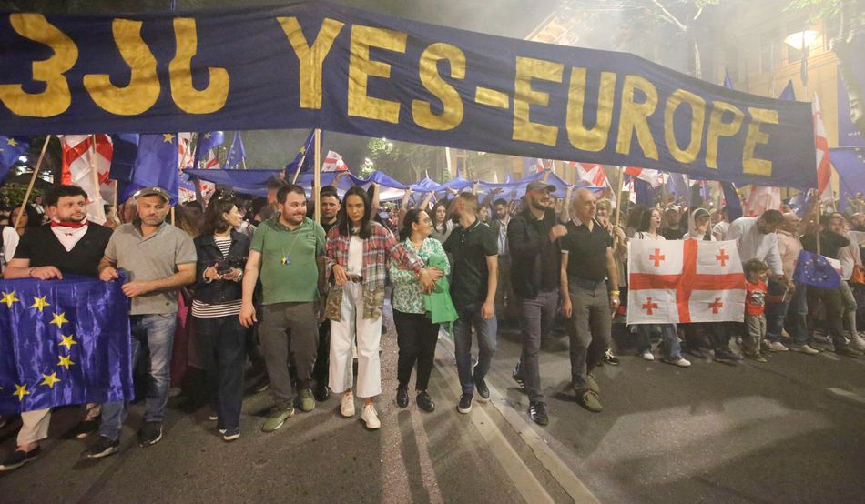 Demonstrators in Tbilisi march under a blue banner with yellow text that reads "Yes-Europe". In the front row, some carry an EU (blue with yellow stars) and a Georgian (white with a large red cross and four smaller red cross) flag. © Associated Press / Alamy