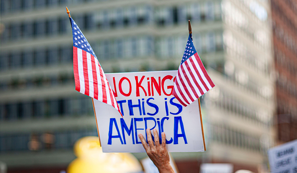 A white placard, held aloft in one hand, reads “No Kings, this is America” in red and blue writing. Two US flags are attached to the placard. Image: Glasshouse Images